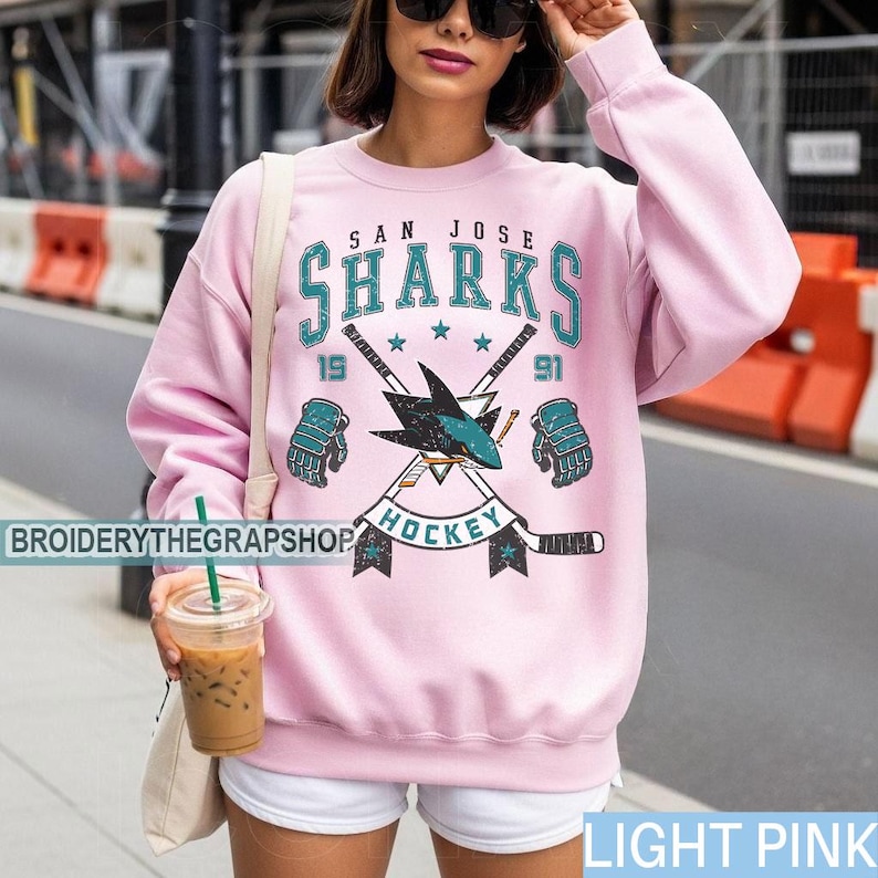 Puede incluir: Una sudadera de cuello redondo rosa claro con un estampado gr&aacute;fico del logotipo de los San Jose Sharks con palos de hockey cruzados, guantes y el texto San Jose Sharks 1991 Hockey. El dise&ntilde;o utiliza colores verde azulado, negro y blanco.