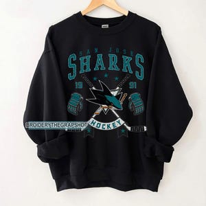 Puede incluir: Una sudadera negra de cuello redondo con un gr&aacute;fico de los San Jose Sharks en color verde azulado y blanco. El dise&ntilde;o incluye el nombre del equipo, el a&ntilde;o 1991, palos de hockey cruzados, dos guantes de hockey y la palabra HOCKEY. El texto BROIDERYTHEGRAPSHOP es visible.