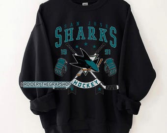 May include: A black crewneck sweatshirt featuring a San Jose Sharks graphic in teal and white. The design includes the team name, the year 1991, crossed hockey sticks, two hockey gloves, and the word HOCKEY. The text BROIDERYTHEGRAPSHOP is visible.