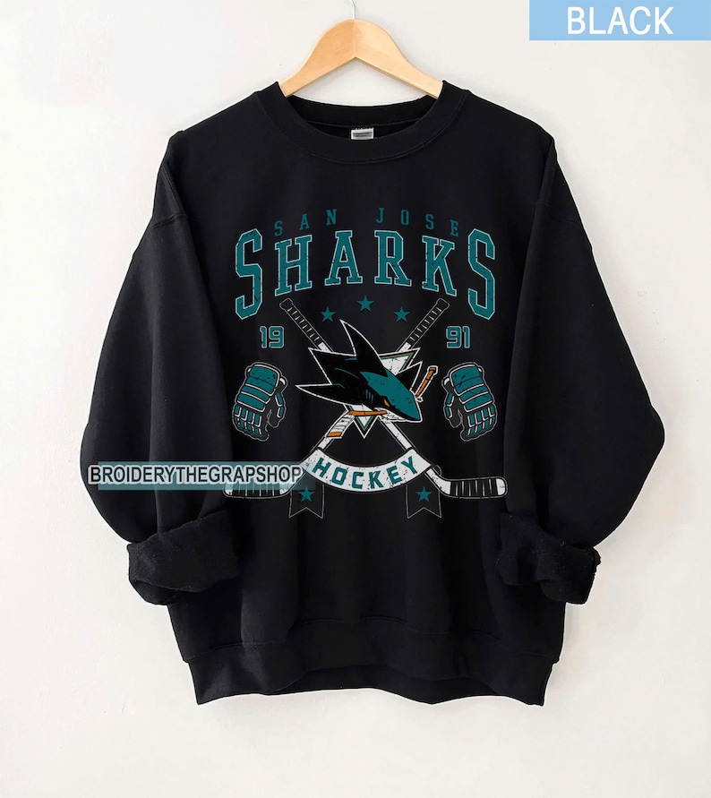 Puede incluir: Una sudadera negra de cuello redondo con un gr&aacute;fico de los San Jose Sharks en color verde azulado y blanco. El dise&ntilde;o incluye el nombre del equipo, el a&ntilde;o 1991, palos de hockey cruzados, dos guantes de hockey y la palabra HOCKEY. El texto BROIDERYTHEGRAPSHOP es visible.