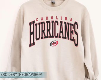 Puede incluir: Una sudadera de cuello redondo color arena con el texto CAROLINA HURRICANES en una fuente negra arqueada y llamativa con contornos rojos. Debajo del texto hay un pequeño logotipo circular de huracán en rojo, negro y blanco en una percha.