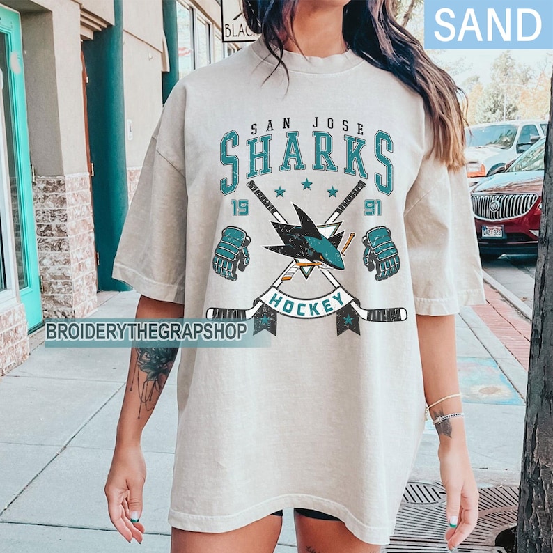 Puede incluir: Una camiseta color arena con un gr&aacute;fico de estilo vintage de los San Jose Sharks. El dise&ntilde;o incluye el texto 'SAN JOSE SHARKS', '1991' y 'HOCKEY', con un logotipo central de tibur&oacute;n, palos de hockey cruzados y guantes en verde azulado y negro.