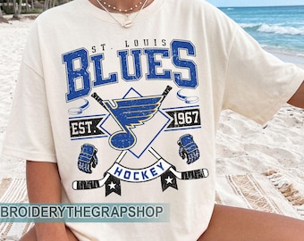 May include: An ivory short-sleeve t-shirt featuring a distressed blue and black graphic print that reads ST. LOUIS BLUES, EST. 1967, and HOCKEY. The design includes a central winged musical note, crossed hockey sticks, gloves, and pucks.