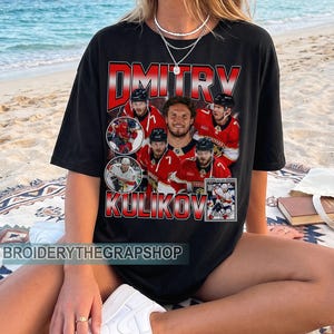 May include: A black short-sleeve t-shirt featuring a graphic print of hockey player Dmitry Kulikov in various action poses and a portrait. The text DMITRY KULIKOV is printed in large red and white letters. The shirt also includes a small rectangular image.