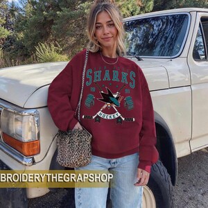 Puede incluir: Una sudadera granate de manga larga con un gr&aacute;fico de hockey de los San Jose Sharks, texto en turquesa y blanco, palos de hockey cruzados y guantes. El dise&ntilde;o incluye los n&uacute;meros 19 y 91. El texto BROIDERYTHEGRAPSHOP es visible en la parte inferior.