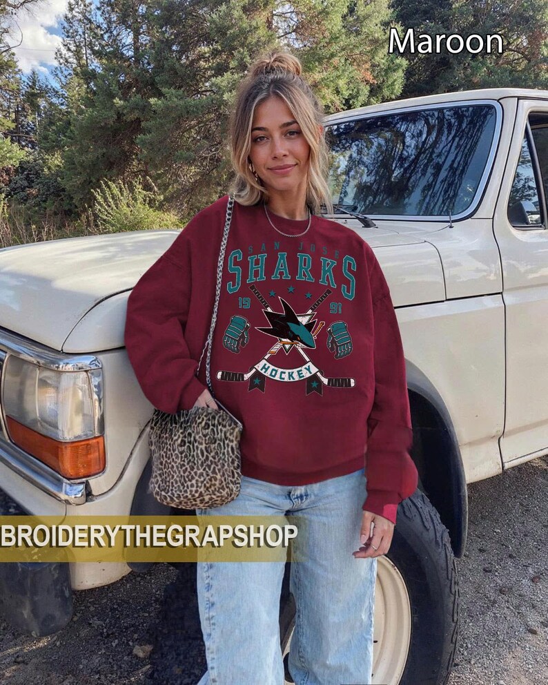 Puede incluir: Una sudadera granate de manga larga con un gr&aacute;fico de hockey de los San Jose Sharks, texto en turquesa y blanco, palos de hockey cruzados y guantes. El dise&ntilde;o incluye los n&uacute;meros 19 y 91. El texto BROIDERYTHEGRAPSHOP es visible en la parte inferior.