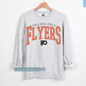 Może przedstawiać: Jasnoszara bluza z okrągłym dekoltem z napisem PHILADELPHIA FLYERS dużymi pomarańczowymi literami z czarnym obrysem. Pod tekstem znajduje się czarno-pomarańczowe logo Philadelphia Flyers. Słowo ASH znajduje się w prawym górnym rogu.