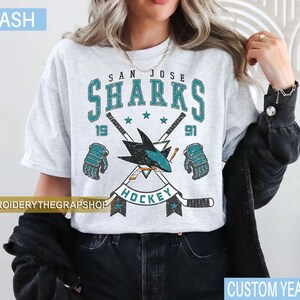 Puede incluir: Una camiseta gris ceniza con un gr&aacute;fico desgastado del equipo de hockey San Jose Sharks. El dise&ntilde;o incluye el texto San Jose Sharks, el a&ntilde;o 1991, dos palos de hockey cruzados, un logotipo de tibur&oacute;n y dos guantes de hockey en turquesa y negro.