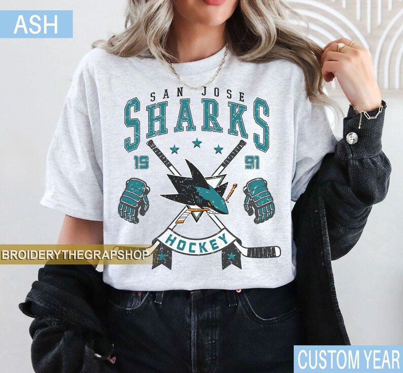 Puede incluir: Una camiseta gris ceniza con un gr&aacute;fico desgastado del equipo de hockey San Jose Sharks. El dise&ntilde;o incluye el texto San Jose Sharks, el a&ntilde;o 1991, dos palos de hockey cruzados, un logotipo de tibur&oacute;n y dos guantes de hockey en turquesa y negro.