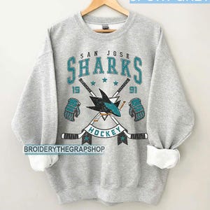 Puede incluir: Una sudadera gris deportiva de cuello redondo con un gr&aacute;fico desgastado del logotipo de los San Jose Sharks, palos de hockey cruzados, guantes y el texto San Jose Sharks Hockey 1991. El dise&ntilde;o utiliza colores verde azulado, negro y blanco.