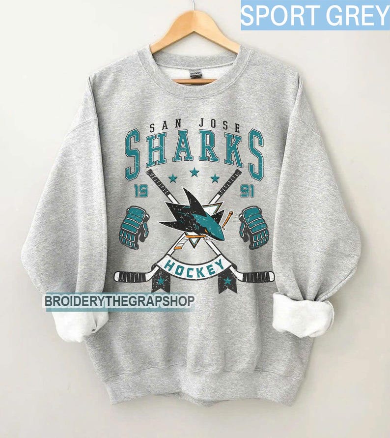Puede incluir: Una sudadera gris deportiva de cuello redondo con un gr&aacute;fico desgastado del logotipo de los San Jose Sharks, palos de hockey cruzados, guantes y el texto San Jose Sharks Hockey 1991. El dise&ntilde;o utiliza colores verde azulado, negro y blanco.