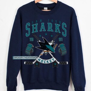 Puede incluir: Una sudadera azul marino de cuello redondo con un estampado gr&aacute;fico del logotipo de los San Jose Sharks, dos palos de hockey cruzados y guantes de hockey. El texto dice SAN JOSE SHARKS, 1991 y HOCKEY. El dise&ntilde;o incluye detalles en color verde azulado y blanco.