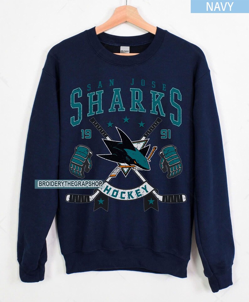 Puede incluir: Una sudadera azul marino de cuello redondo con un estampado gr&aacute;fico del logotipo de los San Jose Sharks, dos palos de hockey cruzados y guantes de hockey. El texto dice SAN JOSE SHARKS, 1991 y HOCKEY. El dise&ntilde;o incluye detalles en color verde azulado y blanco.