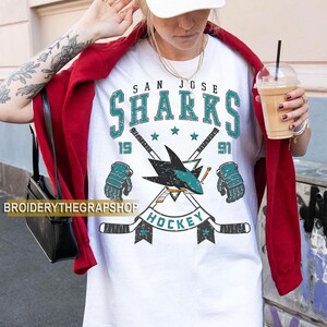 Puede incluir: Una camiseta blanca con un gr&aacute;fico desgastado del equipo de hockey San Jose Sharks. El dise&ntilde;o incluye el texto SAN JOSE SHARKS, el a&ntilde;o 1991, dos palos de hockey cruzados, un logotipo de tibur&oacute;n y dos guantes de hockey en colores verde azulado y negro.