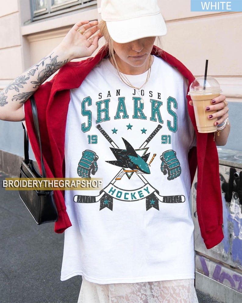 Puede incluir: Una camiseta blanca con un gr&aacute;fico desgastado del equipo de hockey San Jose Sharks. El dise&ntilde;o incluye el texto SAN JOSE SHARKS, el a&ntilde;o 1991, dos palos de hockey cruzados, un logotipo de tibur&oacute;n y dos guantes de hockey en colores verde azulado y negro.
