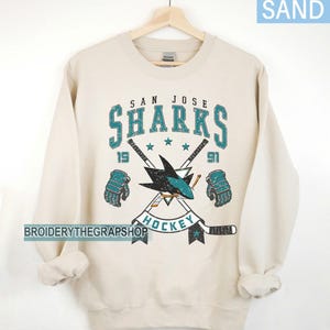 Puede incluir: Una sudadera de cuello redondo color arena con un gr&aacute;fico desgastado del logotipo de los San Jose Sharks. El dise&ntilde;o incluye el texto 'SAN JOSE SHARKS', '1991' y 'HOCKEY', junto con ilustraciones de dos palos de hockey cruzados y dos guantes.