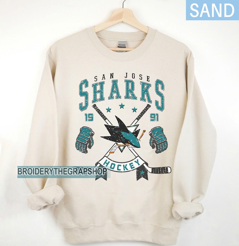 Puede incluir: Una sudadera de cuello redondo color arena con un gr&aacute;fico desgastado del logotipo de los San Jose Sharks. El dise&ntilde;o incluye el texto 'SAN JOSE SHARKS', '1991' y 'HOCKEY', junto con ilustraciones de dos palos de hockey cruzados y dos guantes.
