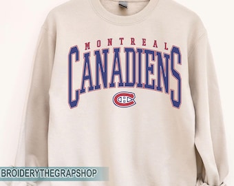 Puede incluir: Una sudadera de cuello redondo color arena que presenta el texto MONTREAL CANADIENS en una fuente azul grande y arqueada con contornos rojos. Debajo del texto se encuentra el logotipo oficial de los Montreal Canadiens en rojo, blanco y azul.