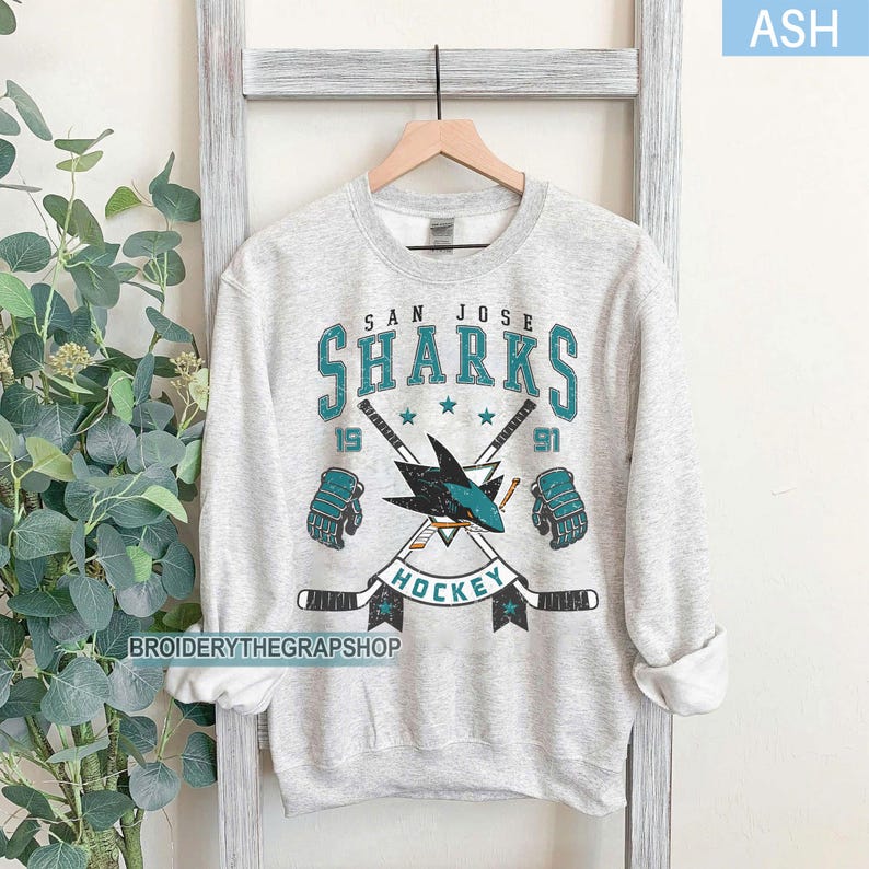 Puede incluir: Una sudadera de cuello redondo gris ceniza con un estampado gr&aacute;fico del logotipo de los San Jose Sharks con palos de hockey cruzados, guantes y el texto San Jose Sharks Hockey 1991. El dise&ntilde;o utiliza colores verde azulado, negro y blanco.