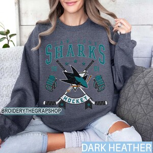 Puede incluir: Una sudadera de cuello redondo en gris jaspeado oscuro con un gr&aacute;fico en color verde azulado y blanco del logotipo de los San Jose Sharks, palos de hockey cruzados y guantes. El texto dice San Jose Sharks 1991 Hockey en el centro del pecho.
