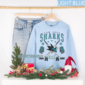 Puede incluir: Una sudadera de cuello redondo azul claro con un gr&aacute;fico de hockey de los San Jose Sharks que incluye palos cruzados, guantes y el texto San Jose Sharks 1991 Hockey. La sudadera se muestra en una percha de madera junto a unos vaqueros claros.