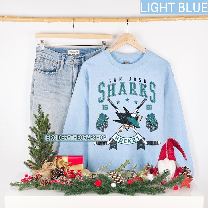 Puede incluir: Una sudadera de cuello redondo azul claro con un gr&aacute;fico de hockey de los San Jose Sharks que incluye palos cruzados, guantes y el texto San Jose Sharks 1991 Hockey. La sudadera se muestra en una percha de madera junto a unos vaqueros claros.