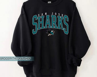 Puede incluir: Una sudadera negra de cuello redondo con el texto SAN JOSE SHARKS en una fuente arqueada de color verde azulado y blanco. Debajo del texto hay un gráfico de un tiburón mordiendo un palo de hockey. Mangas largas con puños y dobladillo acanalados.