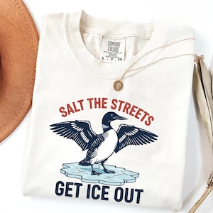 Może przedstawiać: Kremowa koszulka z grafiką nurka z rozłożonymi skrzydłami na krze lodowej. Tekst "SALT THE STREETS" znajduje się nad ptakiem, a "GET ICE OUT" pod nim. Widoczny jest również złoty naszyjnik i brązowy kapelusz.