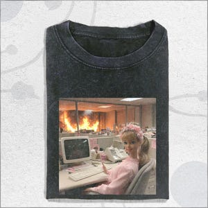 May include: Dark gray t-shirt featuring a doll wearing a pink outfit and a floral crown, seated at a desk with a computer. The background shows an office engulfed in flames. The shirt has a crew neck.