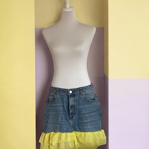Peut inclure: Jupe en jean bleu clair avec un ourlet à volants jaune vif, présentée sur un mannequin. La jupe présente une conception classique à cinq poches. La volants jaune ajoute un contraste ludique au denim.