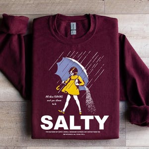 Op de afbeelding: Bordeaux sweatshirt met een afbeelding van een persoon onder een paraplu, zout uitgietend. De persoon draagt een gele jurk en de paraplu is blauw. Het woord "SALTY" staat in het wit onder de afbeelding. Boven de afbeelding staat de tekst "All these flavors and you choose to be".