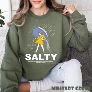 Op de afbeelding: Olijfgroene sweatshirt met het woord "SALTY" in grote witte letters. De afbeelding toont een persoon in een gele jurk onder een blauwe paraplu, zout strooiend. Tekst boven de afbeelding luidt "All the flavors and you choose to be".