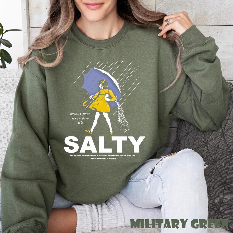 Op de afbeelding: Olijfgroene sweatshirt met het woord "SALTY" in grote witte letters. De afbeelding toont een persoon in een gele jurk onder een blauwe paraplu, zout strooiend. Tekst boven de afbeelding luidt "All the flavors and you choose to be".