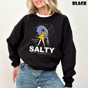 Op de afbeelding: Zwart sweatshirt met een afbeelding van een persoon onder een paraplu, zout strooiend. Het woord "SALTY" staat in grote witte letters. De tekst boven de afbeelding luidt "All the flavors and you choose to be."