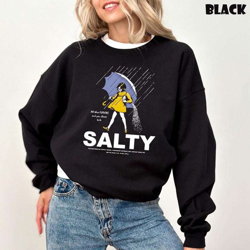 Op de afbeelding: Zwart sweatshirt met een afbeelding van een persoon onder een paraplu, zout strooiend. Het woord "SALTY" staat in grote witte letters. De tekst boven de afbeelding luidt "All the flavors and you choose to be."