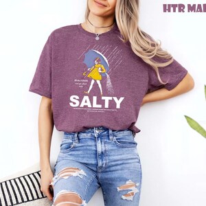 Op de afbeelding: Bordeauxrood t-shirt met een afbeelding van een persoon onder een paraplu, met het woord "SALTY" in het wit. De afbeelding bevat ook de tekst "All the flavors and you chose to be..."