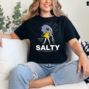 Op de afbeelding: Zwart t-shirt met een afbeelding van een persoon onder een paraplu, zout strooiend. Het woord "SALTY" staat in grote witte letters onder de illustratie. De tekst "All the flavors and you choose to be" staat erboven.