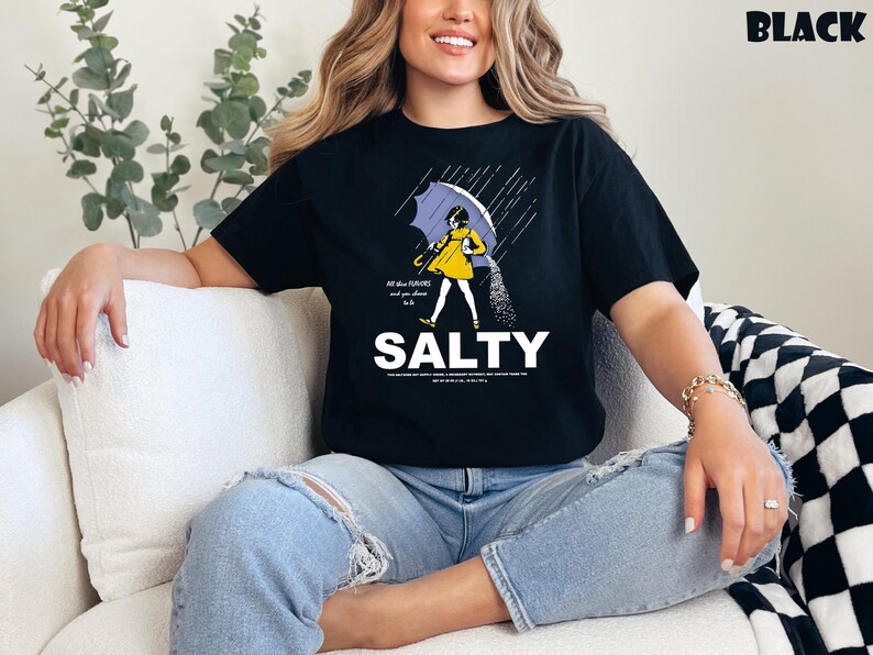 Op de afbeelding: Zwart t-shirt met een afbeelding van een persoon onder een paraplu, zout strooiend. Het woord "SALTY" staat in grote witte letters onder de illustratie. De tekst "All the flavors and you choose to be" staat erboven.