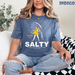 Op de afbeelding: Indigo blauw t-shirt met een afbeelding van een persoon onder een paraplu, zout strooiend. Het woord "SALTY" is gedrukt in grote witte letters. Het shirt heeft de tekst "All the flavors and you choose to be".