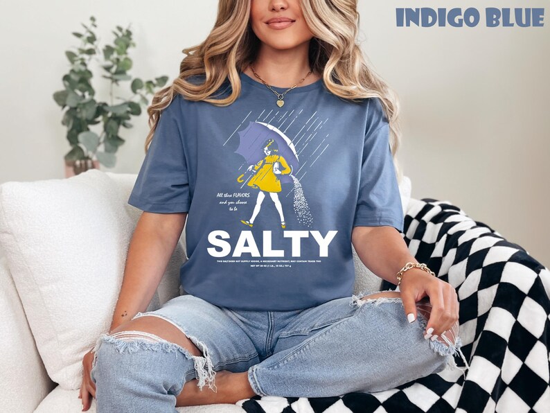 Op de afbeelding: Indigo blauw t-shirt met een afbeelding van een persoon onder een paraplu, zout strooiend. Het woord "SALTY" is gedrukt in grote witte letters. Het shirt heeft de tekst "All the flavors and you choose to be".