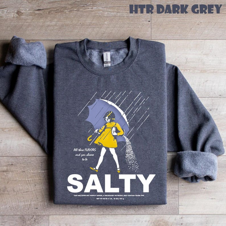 Op de afbeelding: Donkergrijze sweatshirt met het woord "SALTY" in het wit. De afbeelding toont een persoon in een gele jurk onder een blauwe paraplu, met zout dat naar beneden valt. De tekst "HTR DARK GREY" staat bovenaan.