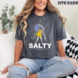 Op de afbeelding: Donkergrijs t-shirt met een afbeelding van een persoon die in de regen loopt onder een paraplu, zout strooiend. Het woord "SALTY" is in grote witte letters onder de afbeelding gedrukt. Het shirt heeft ook de tekst "All the flavors and you choose to be".