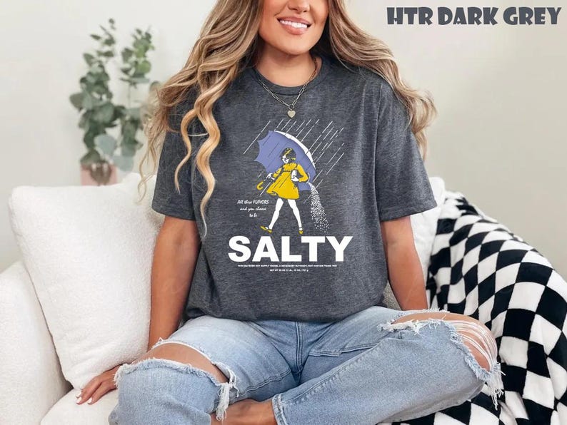Op de afbeelding: Donkergrijs t-shirt met een afbeelding van een persoon die in de regen loopt onder een paraplu, zout strooiend. Het woord "SALTY" is in grote witte letters onder de afbeelding gedrukt. Het shirt heeft ook de tekst "All the flavors and you choose to be".