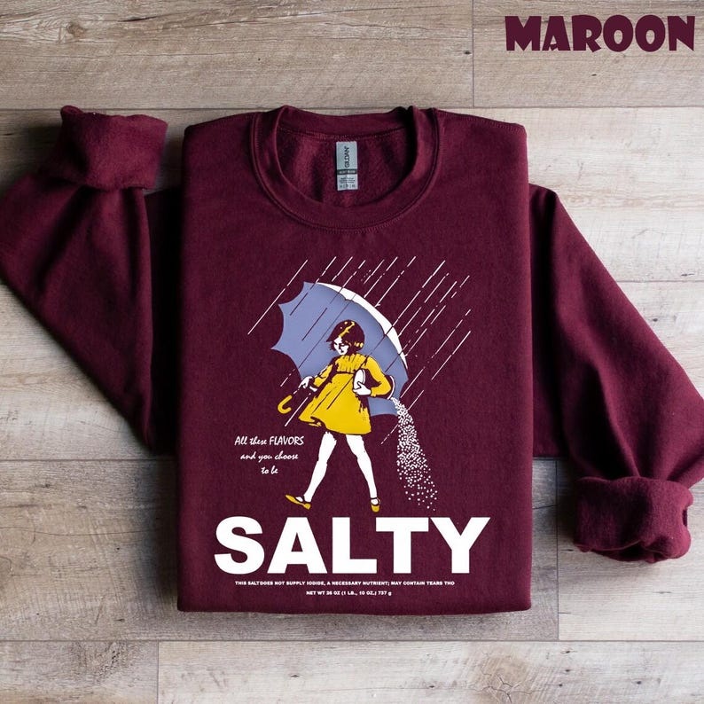 Op de afbeelding: Bordeaux sweatshirt met een afbeelding van een persoon onder een paraplu, zout uitgietend. De persoon draagt een gele jurk en de paraplu is blauw. Het woord "SALTY" staat in het wit onder de afbeelding. Boven de afbeelding staat de tekst "All these flavors and you choose to be".