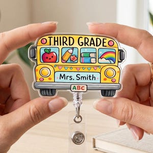 Può includere: Un porta badge a forma di scuolabus giallo con il testo THIRD GRADE e Mrs. Smith. La grafica del bus include illustrazioni di una mela rossa, una matita, libri e un arcobaleno. Una piccola targa in basso mostra le lettere ABC in un riquadro.