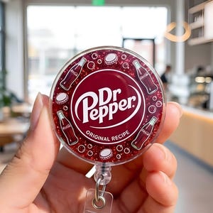 May include: A round, burgundy Dr. Pepper badge reel with the brand's logo in white. The design includes illustrations of Dr. Pepper bottles and bottle caps. The reel has a clear plastic strap and a metal clip.