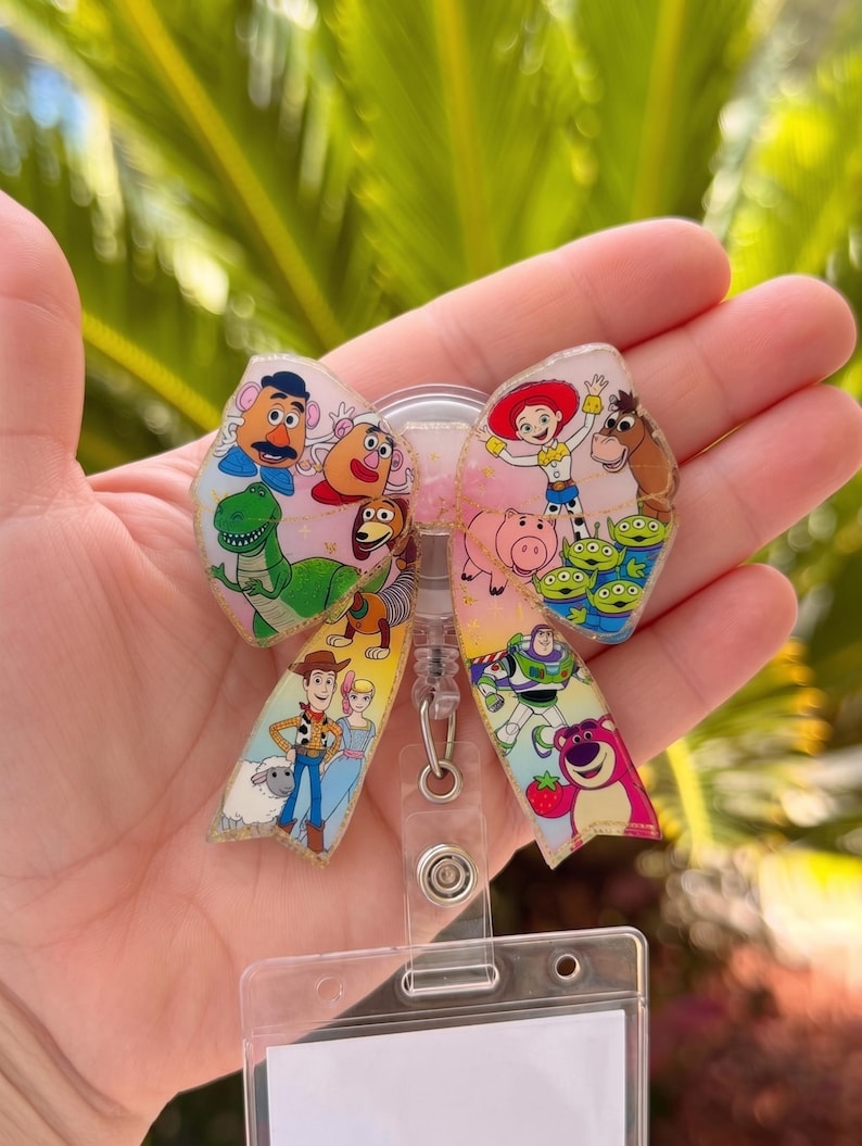 May include: A badge reel featuring a bow-shaped acrylic topper decorated with colourful illustrations of various animated toy characters, including a cowboy, a space ranger, a cowgirl, a bear, and several aliens, attached to a clear plastic card holder.