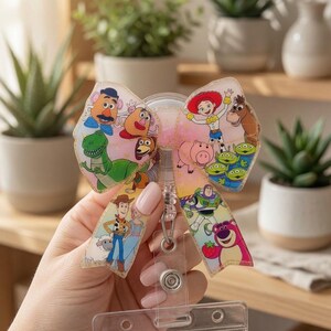 May include: A badge reel featuring a pink glittery bow adorned with various Toy Story character illustrations including Woody, Buzz Lightyear, Jessie, Rex, Mr. Potato Head, and the Aliens. The bow is attached to a clear plastic ID card holder.