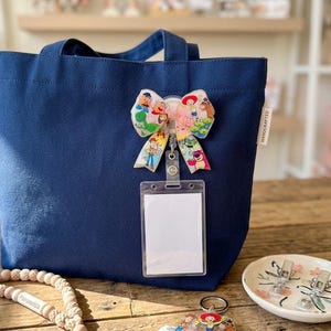 May include: A navy blue canvas tote bag featuring a handcrafted bow badge with a Toy Story character print, attached to a clear plastic ID card holder. A matching character keychain and a wooden bead lanyard are placed on the wooden surface.