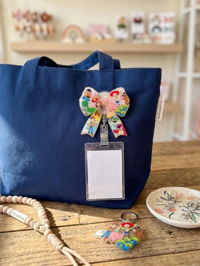 May include: A navy blue canvas tote bag featuring a handcrafted bow badge with a Toy Story character print, attached to a clear plastic ID card holder. A matching character keychain and a wooden bead lanyard are placed on the wooden surface.